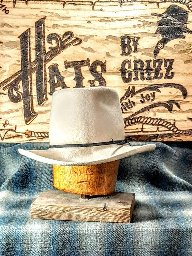 Puede incluir: Un sombrero vaquero de fieltro blanco con una banda negra, exhibido en un soporte de sombrero de madera. El sombrero est&aacute; delante de un letrero de madera con el texto "Hats By Grizz with Joy".