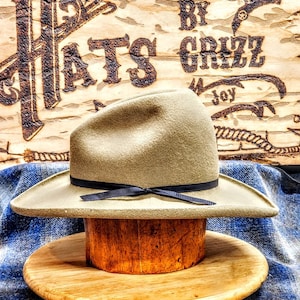 Puede incluir: Un sombrero vaquero marr&oacute;n claro con una cinta negra y un soporte de sombrero de madera. El sombrero est&aacute; sentado sobre una tela a cuadros azul y blanca. El fondo es un letrero de madera con el texto "Hats By Grizz Joy".