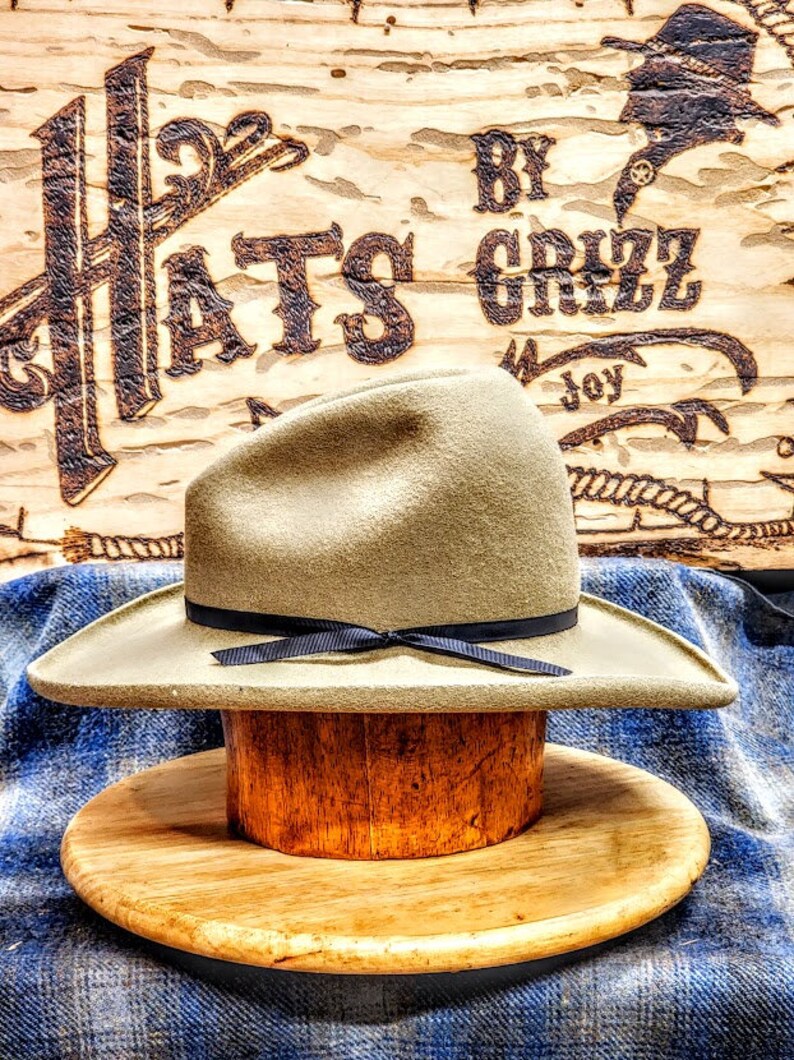 Puede incluir: Un sombrero vaquero marr&oacute;n claro con una cinta negra y un soporte de sombrero de madera. El sombrero est&aacute; sentado sobre una tela a cuadros azul y blanca. El fondo es un letrero de madera con el texto "Hats By Grizz Joy".