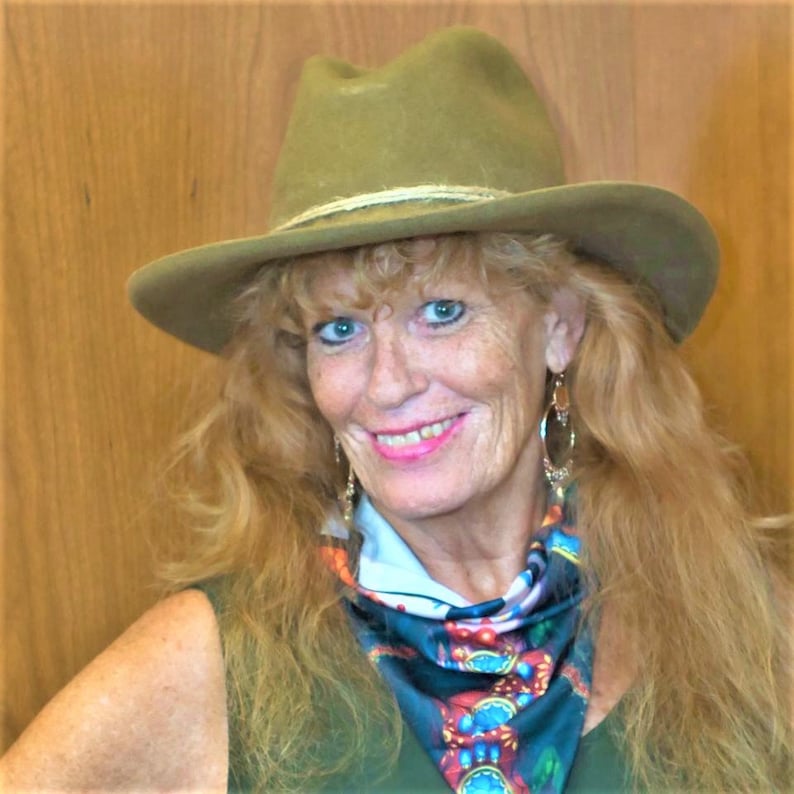 Puede incluir: Una mujer con el pelo largo y rubio que lleva un sombrero verde, un top verde sin mangas y una bufanda colorida. Est&aacute; sonriendo y mirando a la c&aacute;mara.