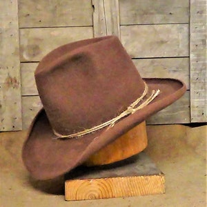 Puede incluir: Un sombrero vaquero de fieltro marr&oacute;n con un ala ancha y una banda de cuero marr&oacute;n. El sombrero est&aacute; sentado en un soporte de madera.