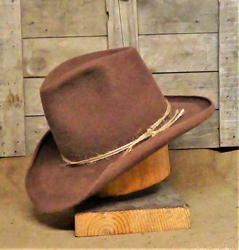 Puede incluir: Un sombrero vaquero de fieltro marr&oacute;n con un ala ancha y una banda de cuero marr&oacute;n. El sombrero est&aacute; sentado en un soporte de madera.