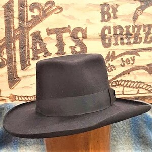 Może przedstawiać: Czarny filcowy kapelusz fedora z szerokim rondem i czarną wstążką grosgrain. Kapelusz leży na drewnianej powierzchni z tabliczką z napisem "Hats by Grizz with Joy".