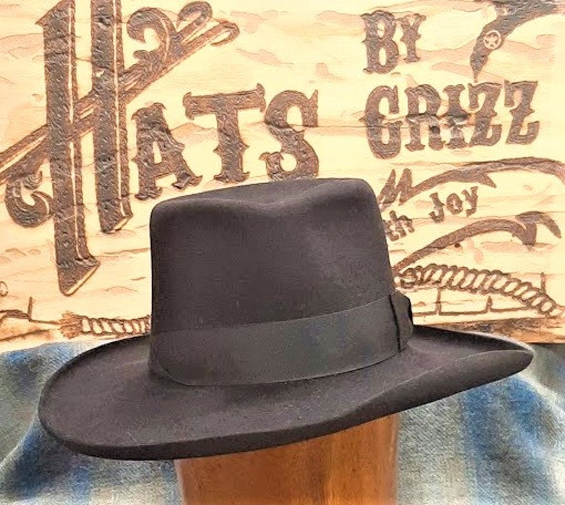 Może przedstawiać: Czarny filcowy kapelusz fedora z szerokim rondem i czarną wstążką grosgrain. Kapelusz leży na drewnianej powierzchni z tabliczką z napisem "Hats by Grizz with Joy".