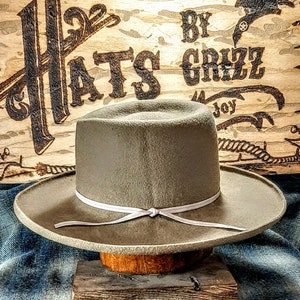 Może przedstawiać: Jasny szary filcowy kapelusz kowbojski z białym sk&oacute;rzanym paskiem i węzłem. Kapelusz stoi na drewnianym stojaku. Tło to drewniany znak z napisem "Hats By Grizz".
