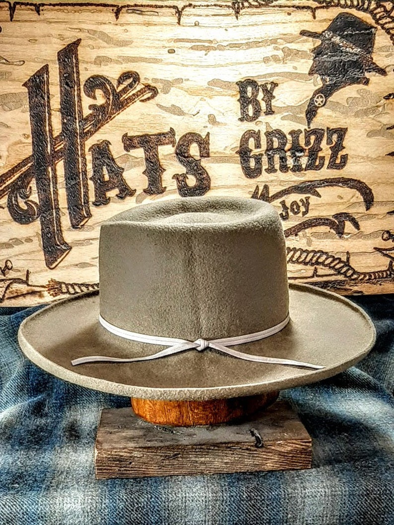 Może przedstawiać: Jasny szary filcowy kapelusz kowbojski z białym sk&oacute;rzanym paskiem i węzłem. Kapelusz stoi na drewnianym stojaku. Tło to drewniany znak z napisem "Hats By Grizz".
