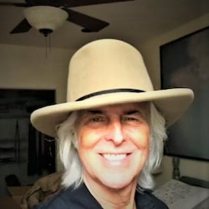 Puede incluir: Un hombre con el pelo largo y blanco lleva un sombrero fedora de fieltro beige con una banda negra. Est&aacute; sonriendo y mirando a la c&aacute;mara.