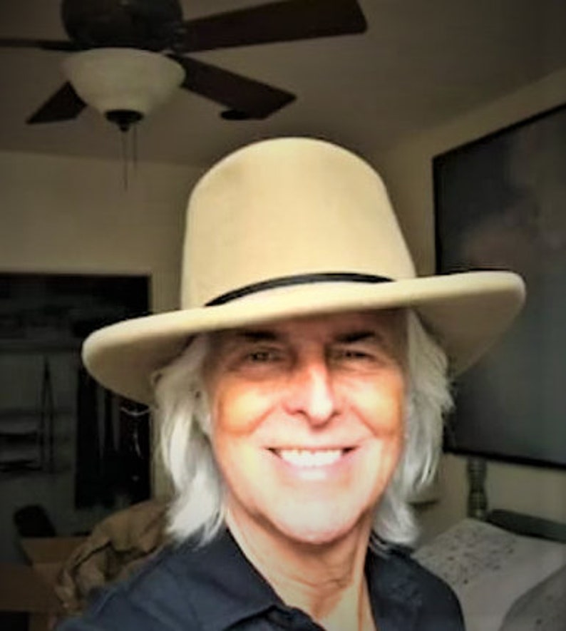 Puede incluir: Un hombre con el pelo largo y blanco lleva un sombrero fedora de fieltro beige con una banda negra. Est&aacute; sonriendo y mirando a la c&aacute;mara.