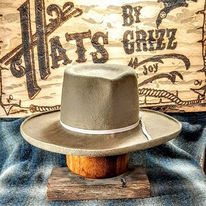Może przedstawiać: Jasny szary filcowy kapelusz kowbojski z białym pasem. Kapelusz stoi na drewnianym bloku. Kapelusz znajduje się przed drewnianym znakiem z napisem "Hats by Grizz".