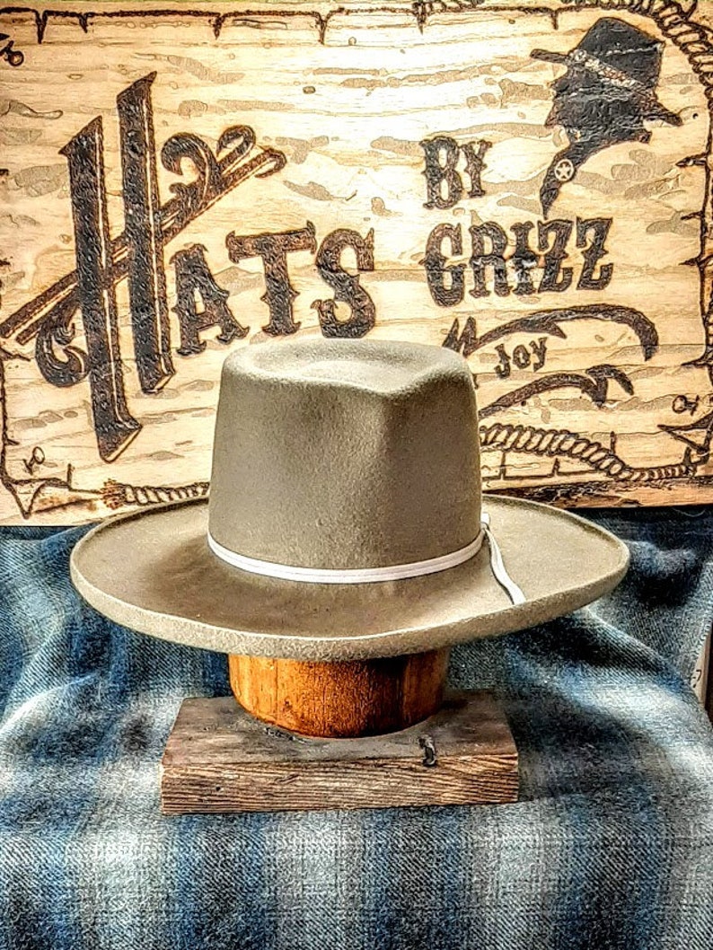 Może przedstawiać: Jasny szary filcowy kapelusz kowbojski z białym pasem. Kapelusz stoi na drewnianym bloku. Kapelusz znajduje się przed drewnianym znakiem z napisem "Hats by Grizz".