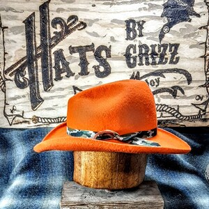 Puede incluir: Sombrero de vaquero de fieltro naranja con una banda de cinta de camuflaje. El sombrero se muestra en un soporte de sombrero de madera. El letrero detr&aacute;s del sombrero dice "Hats By Grizz".