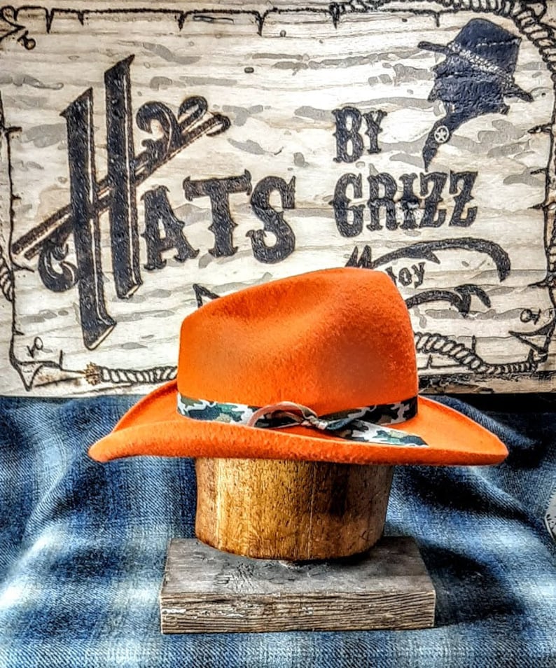 Puede incluir: Sombrero de vaquero de fieltro naranja con una banda de cinta de camuflaje. El sombrero se muestra en un soporte de sombrero de madera. El letrero detr&aacute;s del sombrero dice "Hats By Grizz".