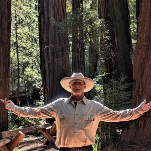 Puede incluir: Una persona con una camisa de botones de color beige y un sombrero blanco se para con los brazos extendidos en un bosque de altos &aacute;rboles de secuoya.
