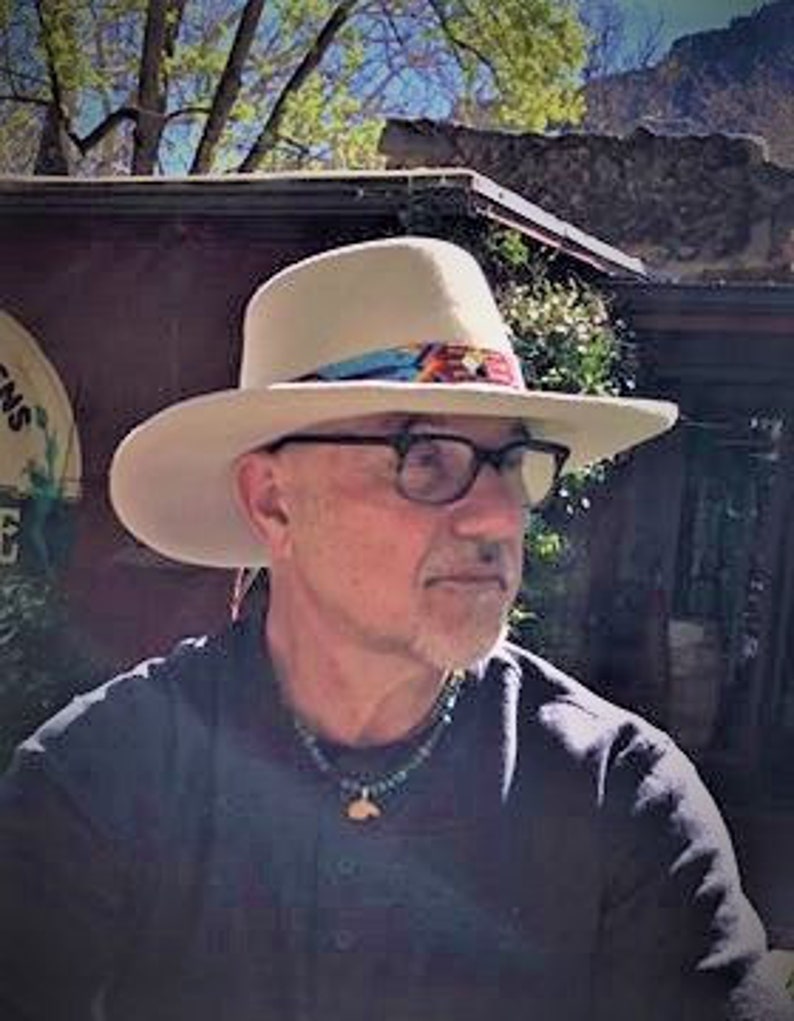 Może przedstawiać: Biały filcowy kapelusz kowbojski z brązowym sk&oacute;rzanym paskiem i kolorową plecioną wstążką. Kapelusz ma szeroki rondel i wysoki korpus.