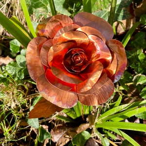 May include: A handcrafted copper rose sculpture with layered petals, displaying a rich, reddish-brown hue. The metal flower is surrounded by green grass and foliage, creating a natural contrast. The sculpture's intricate details are highlighted by sunlight.