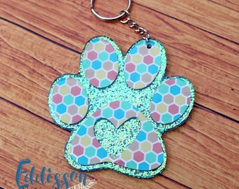 May include: A turquoise blue glitter paw print keychain with a pink, yellow, and blue honeycomb pattern and a small heart in the center.