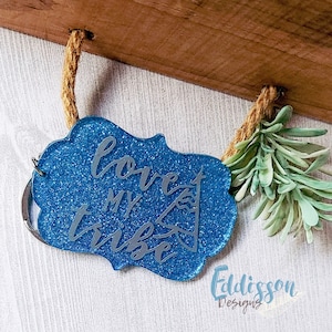 May include: A blue glitter keychain with a silver inscription that reads "love my tribe". The keychain is attached to a piece of twine and a small green plant.