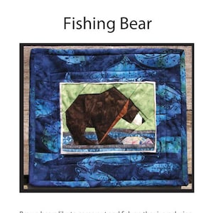 Op de afbeelding: Een patchwork-deken met een afbeelding van een bruine beer die zalm vist in een blauwe en groene rivier. De deken is omlijst met een blauwe stof met een zalmpatroon. De tekst "Fishing Bear" staat bovenaan de afbeelding. De tekst "©Copyright 2015 Maggie Lampton. Ketchikan, Alaska" staat onderaan de afbeelding.