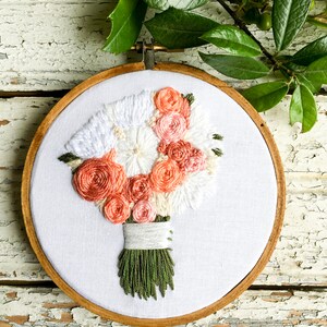 May include: A white embroidery hoop with a hand-stitched bouquet of peach and white flowers. The flowers are arranged in a circular shape and are tied together with a white ribbon. The hoop is made of wood and has a natural finish.