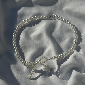 May include: A white pearl necklace with a pearl bow accent. The necklace is on a white fabric background.