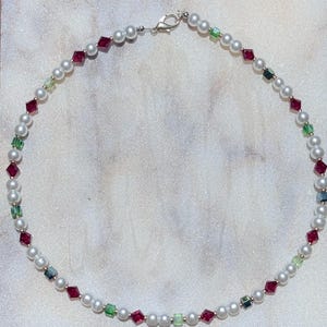 May include: A delicate necklace featuring white pearls interspersed with red, green, and silver-colored crystal beads. The necklace is secured with a silver clasp and is displayed on a white, textured surface.