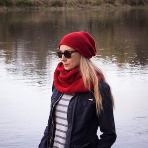 May include: A woman wearing a red knitted hat and scarf, sunglasses, and a navy blue jacket with a striped shirt underneath.