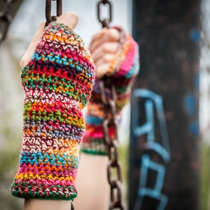 Könnte beinhalten: Ein Paar bunte, fingerlose Häkelhandschuhe mit einem mehrfarbigen Streifenmuster. Die Handschuhe sind aus verschiedenen Garnfarben gefertigt, darunter Rot, Orange, Gelb, Grün, Blau, Lila und Braun.