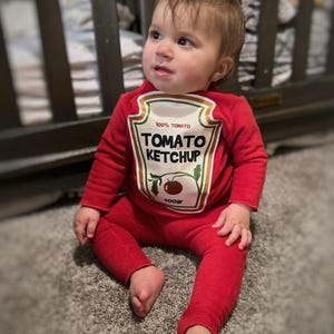 May include: A baby wearing a red Tomato Ketchup costume, with the words "100% Tomato" and "Tomato Ketchup" printed on the front. The baby is sitting on a gray carpet, with a wooden crib in the background.
