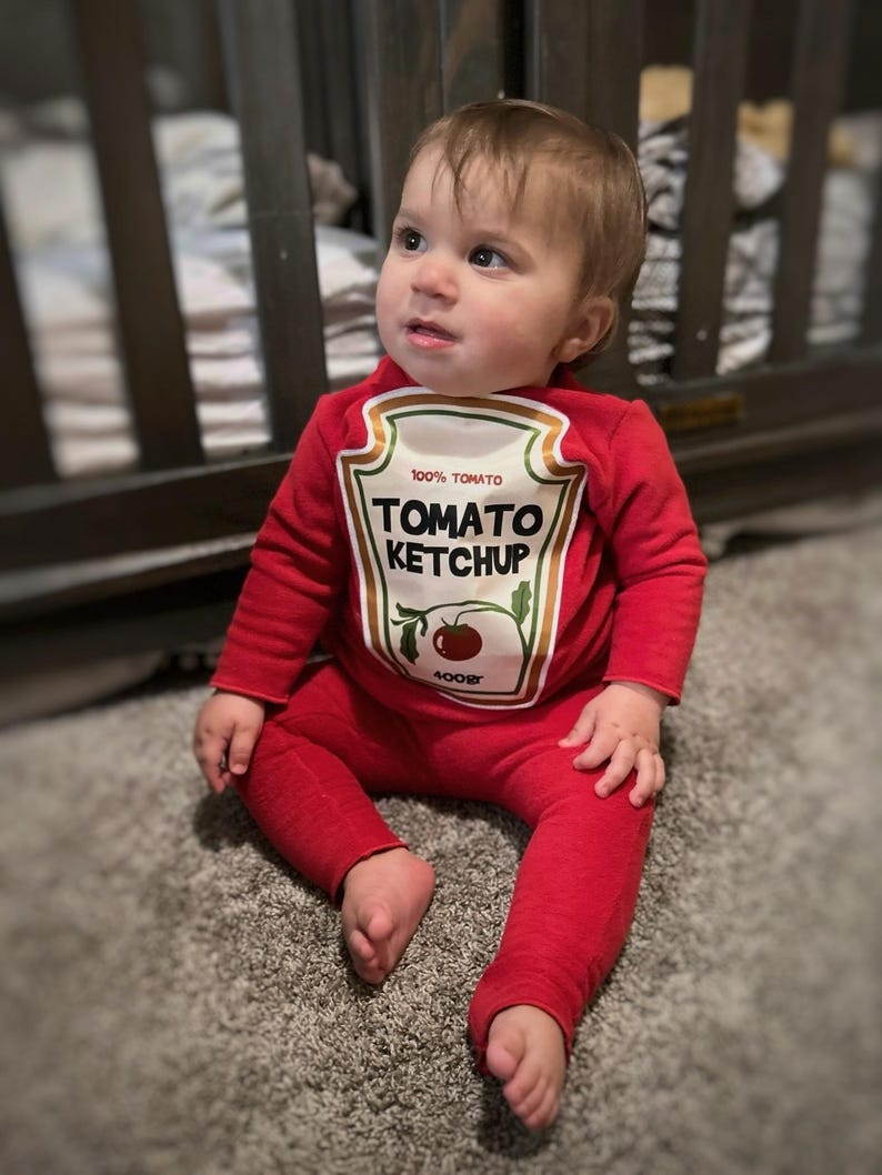 May include: A baby wearing a red Tomato Ketchup costume, with the words "100% Tomato" and "Tomato Ketchup" printed on the front. The baby is sitting on a gray carpet, with a wooden crib in the background.