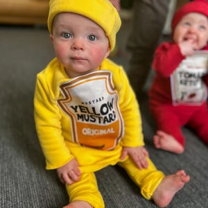May include: A baby dressed in a yellow mustard costume, complete with a matching hat and a tunic printed with a mustard label. The costume includes yellow pants. Another baby in a red tomato costume is in the background.
