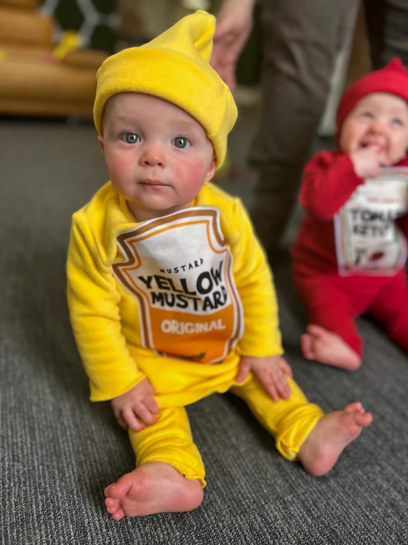 May include: A baby dressed in a yellow mustard costume, complete with a matching hat and a tunic printed with a mustard label. The costume includes yellow pants. Another baby in a red tomato costume is in the background.