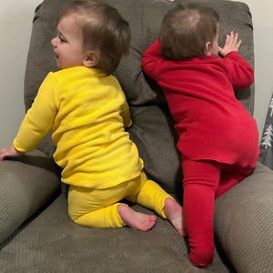 May include: Two babies wearing matching outfits are seated on a gray upholstered chair. One baby is dressed in a yellow long-sleeved top and pants, while the other wears a red outfit. The babies are facing away from the camera.