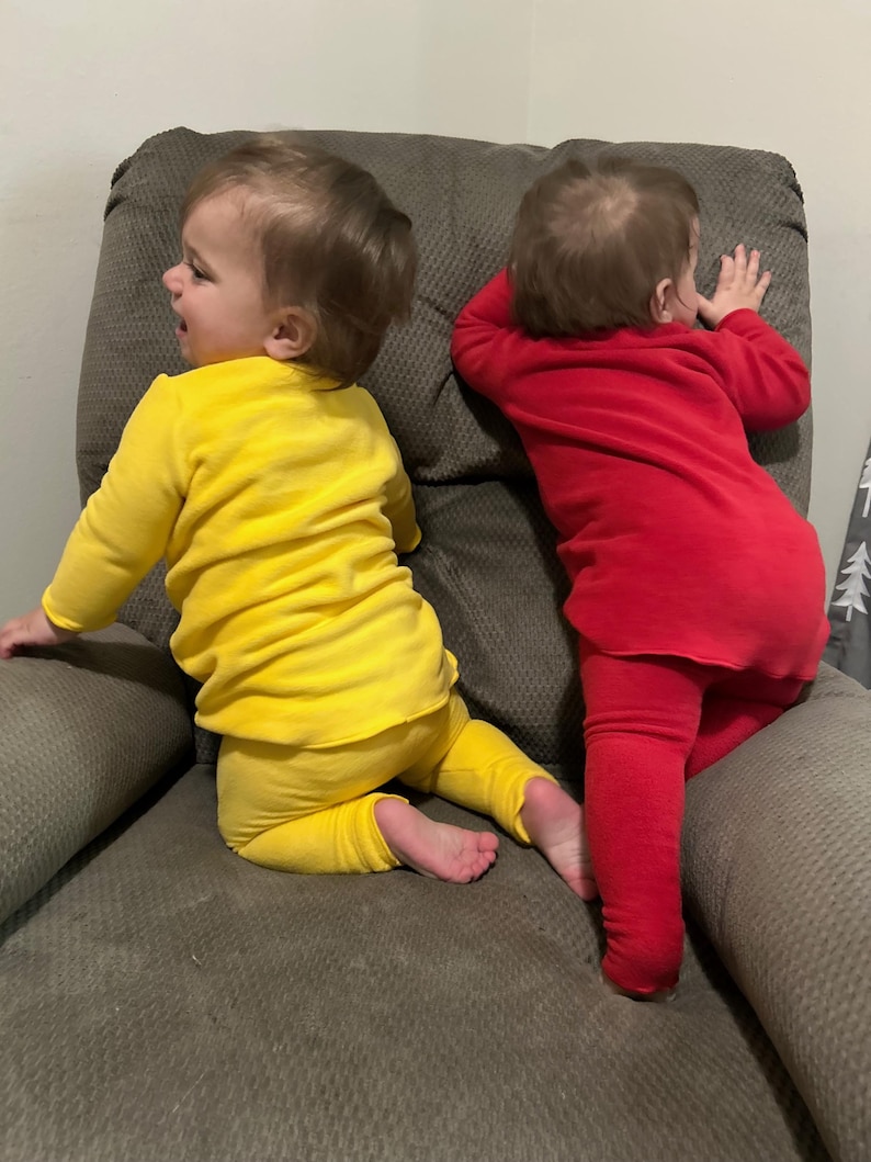 May include: Two babies wearing matching outfits are seated on a gray upholstered chair. One baby is dressed in a yellow long-sleeved top and pants, while the other wears a red outfit. The babies are facing away from the camera.