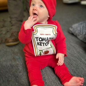 May include: A baby dressed in a red ketchup costume, including a hat and a long-sleeved shirt with a ketchup label design. The baby is sitting on a gray carpet, looking upwards with their mouth open.