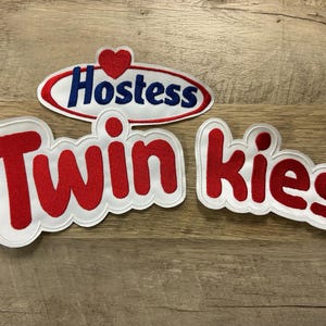 May include: Three embroidered patches with the Hostess and Twinkies logos. The Hostess patch is an oval with red trim, blue text, and a red heart. The Twinkies patches feature red text with white trim.
