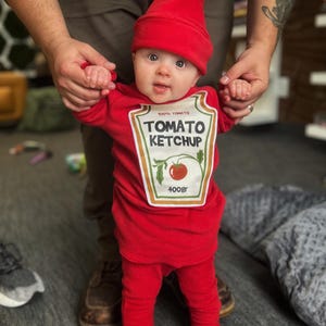 May include: A baby dressed in a red Tomato Ketchup costume, including a matching hat and pants. The costume features a label with the words "Tomato Ketchup" and a tomato illustration. The baby is being held by an adult.