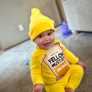 May include: A smiling baby wearing a bright yellow mustard costume, including a hat and a jumpsuit. The costume features a label that reads "Yellow Mustard Original". The baby is sitting on a carpet.