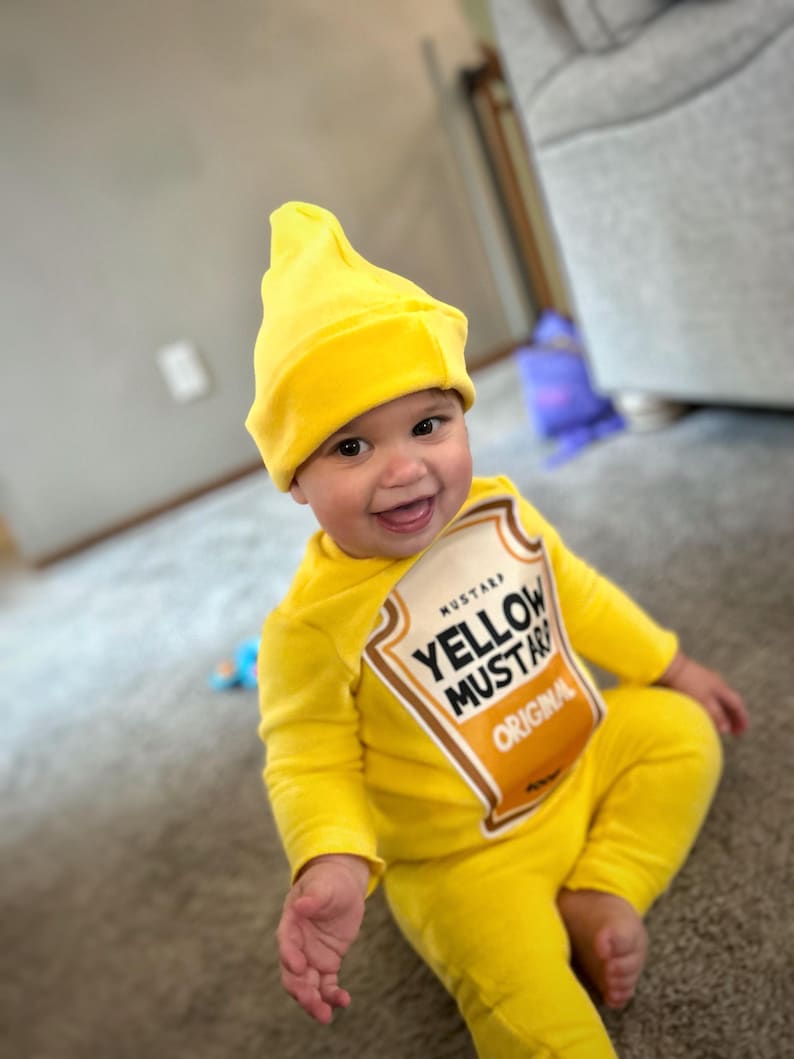 May include: A smiling baby wearing a bright yellow mustard costume, including a hat and a jumpsuit. The costume features a label that reads "Yellow Mustard Original". The baby is sitting on a carpet.