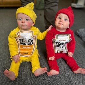 May include: Two babies dressed in food-themed costumes. One wears a yellow mustard outfit with a matching hat, the other a red ketchup costume with a matching hat. The mustard costume has "YELLOW MUSTARD ORIGINAL 400gr" printed on it. The ketchup costume has "100% TOMATO KETCHUP" printed on it.