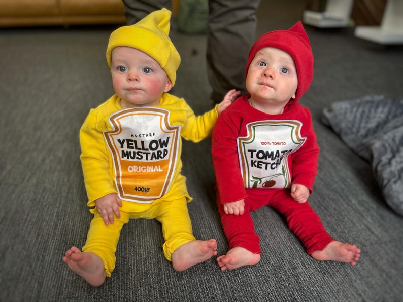 May include: Two babies dressed in food-themed costumes. One wears a yellow mustard outfit with a matching hat, the other a red ketchup costume with a matching hat. The mustard costume has "YELLOW MUSTARD ORIGINAL 400gr" printed on it. The ketchup costume has "100% TOMATO KETCHUP" printed on it.