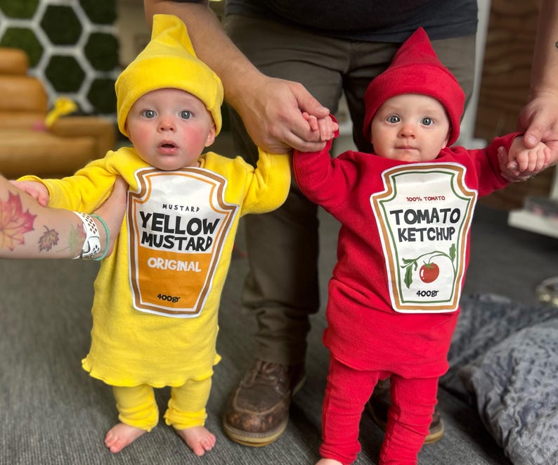 May include: Two babies dressed in food-themed costumes. One wears a yellow mustard costume with a matching hat, the other a red ketchup costume with a matching hat. The mustard costume has the text "YELLOW MUSTARD ORIGINAL 400gr". The ketchup costume has the text "TOMATO KETCHUP 100% TOMATO 400gr".
