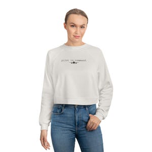May include: A white cropped sweatshirt with long sleeves and the text "pilot in command." in black. The sweatshirt is paired with blue jeans.