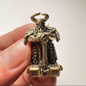 Peut inclure: Une petite figurine métallique aux tons laiton représentant un guerrier barbu portant un casque à cornes et une armure texturée. Le personnage se tient debout, les deux mains posées sur le manche d'un grand marteau à deux têtes.