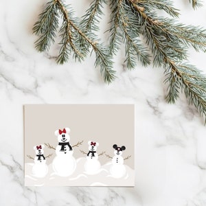 May include: A greeting card featuring four snowmen with mouse ears, black scarves, and twig arms. The snowmen have red or black bows. The card is on a beige background with a marble surface and a sprig of evergreen.