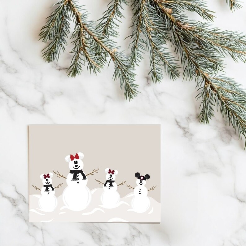 May include: A greeting card featuring four snowmen with mouse ears, black scarves, and twig arms. The snowmen have red or black bows. The card is on a beige background with a marble surface and a sprig of evergreen.
