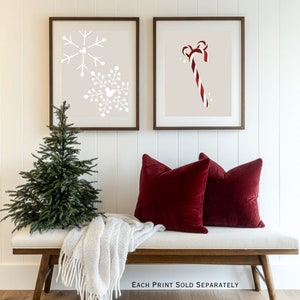 Op de afbeelding: Twee ingelijste prints met een kerstthema. E&eacute;n toont witte sneeuwvlokken op een beige achtergrond, de andere een rood-witte zuurstok. Daaronder twee rode fluwelen kussens op een bank met een witte deken en een kleine kerstboom.