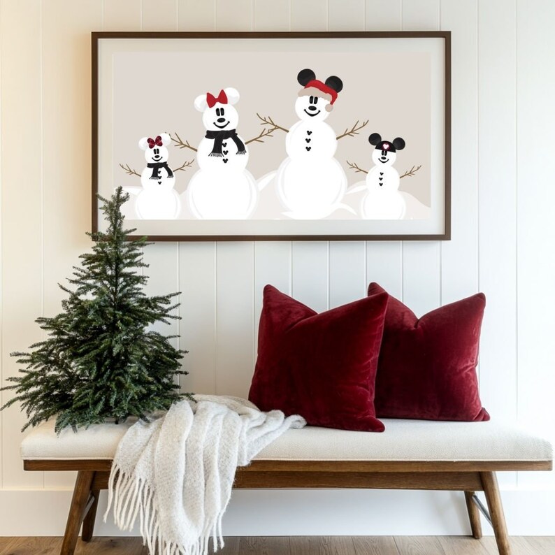 May include: A framed print featuring a family of snowmen with Mickey Mouse ears, scarves, and hats. The print hangs above a bench with red velvet pillows and a gray throw blanket. A small Christmas tree is to the left.