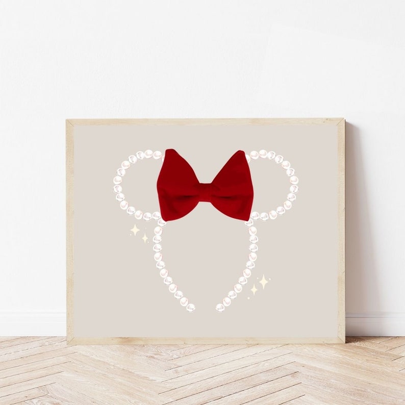 May include: A framed print featuring a red bow and pearl-like beads arranged in the shape of mouse ears. The background is a light beige color, with sparkling accents. The artwork is displayed on a wooden floor.