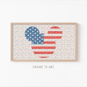 Op de afbeelding: Een ingelijste print van een Mickey Mouse-silhouet met het ontwerp van de Amerikaanse vlag. De achtergrond is een lichtblauw en roze bloemenpatroon. De tekst "FRAME TV ART" staat onder de afbeelding.