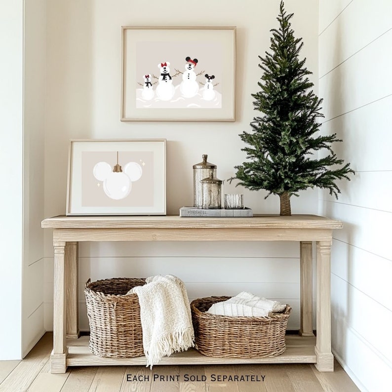 May include: A wooden console table with two framed prints and two woven baskets. One print features a snowman family with Mickey Mouse ears, the other a Mickey Mouse ornament. A small Christmas tree and decorative jars are also on the table.